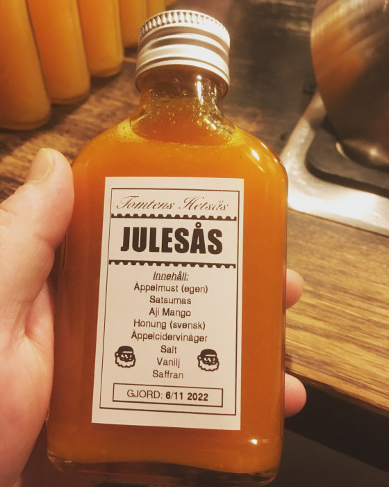 En hand håller en flaska märkt "JULESÅS" med ingredienser som äppelmust och saffran, tillverkad den 6 november 2022.