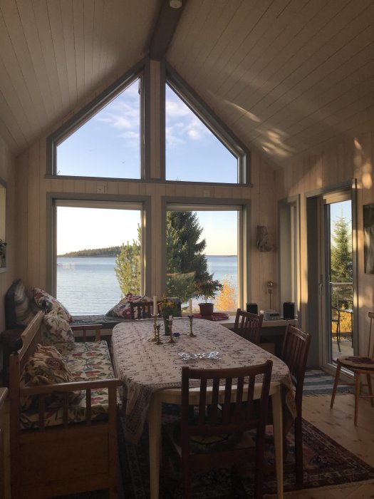 Vardagsrum med matbord, katedralfönster, utsikt över sjö, naturligt ljus, hemtrevlig inredning, terrassdörrar.