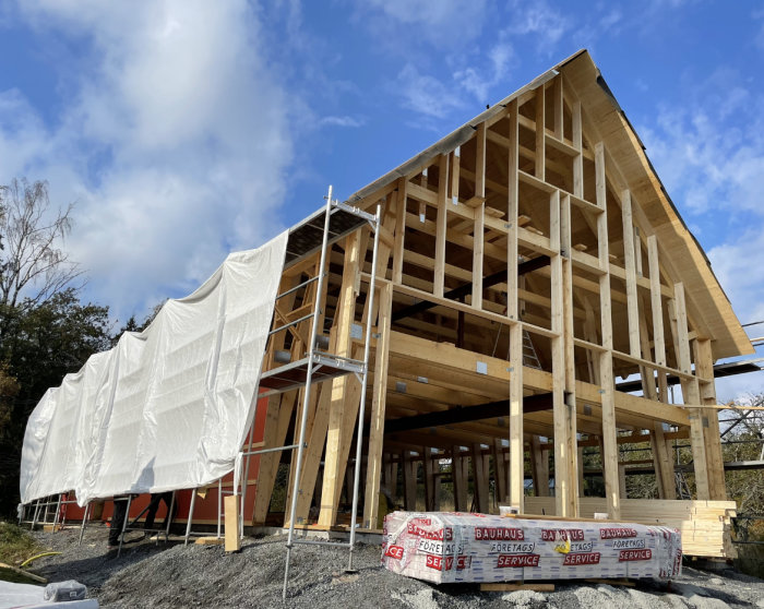 Trästomme för byggnad under konstruktion, täckt delvis med presenning, byggmaterial synligt, molnig himmel.