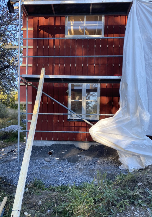 Renovering av rödfärgat hus, byggnadsställning, plastöverdrag, fönster, byggmaterial på marken, utomhus.