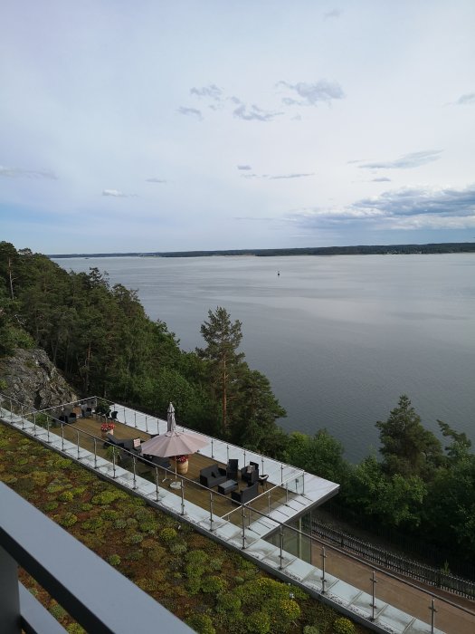 Uteservering på klippa, utsikt över sjö, molnigt, grönskande tak, räcke, natur och lugn atmosfär.