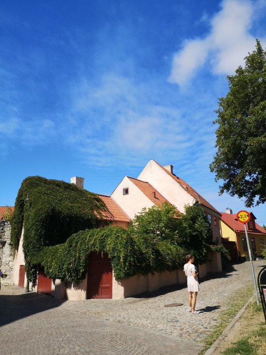 Gammal kullerstensgata, färgglada byggnader, grönska, klarblå himmel, person promenerar, sommar, stadsmiljö.