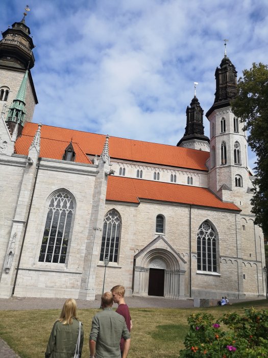 Tre personer promenerar mot en stor, historisk kyrka med dubbla torn under en blå himmel.