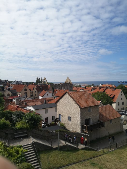 Historisk stadsvy, tegeltak, gammal stenbyggnad, turister, delvis molnig himmel, hav i bakgrunden.