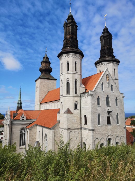 En gammal stenkyrka med två torn mot en blå himmel, omgiven av grönska, synlig delvis hav i bakgrunden.