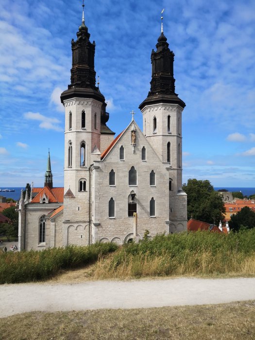 Kyrkobyggnad med två torn, moln på himlen, grönområde framför, synlig stad och vatten i bakgrunden.