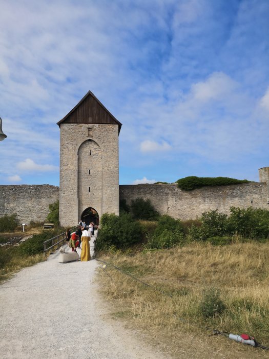 Medeltida borgtorn, stenväggar, personer i historiska kläder, blå himmel, sommardag.