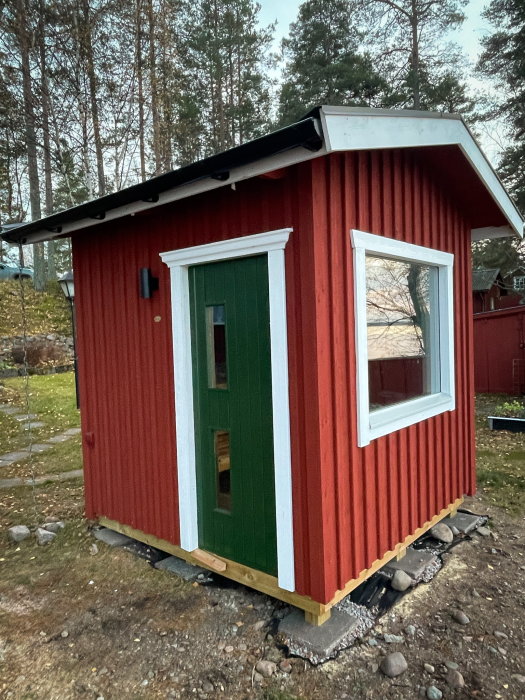 Rött trähus med vita knutar, gröna dörrar och fönster, står i skogsmiljö, stenar runt grunden.