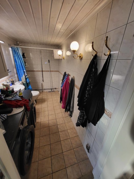 Långsmalt badrum med kläder på krokar, spegel, tvättmaskin och duschdraperi. Kombinerar tvätt med hygien.