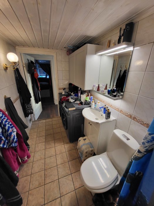 Ett trångt, ostädat badrum med många föremål på golvet och bänkar, inredning i vit och träfärg.