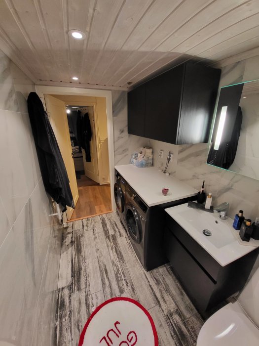 Modernt badrum med tvättmaskin, svart skåp, spegel, vit handfat, trädetaljer på väggar och tak.