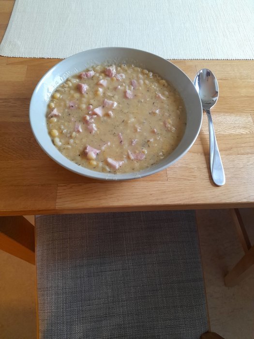 En skål med ärtsoppa och skinkbitar på ett träbord, bredvid en sked på en servett.