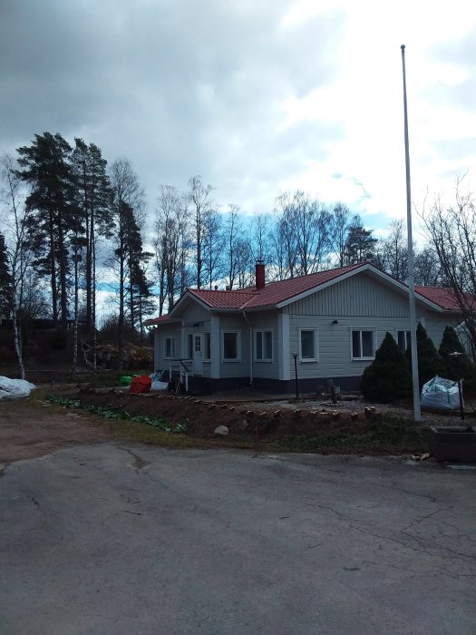 Ett grått hus med röda takpannor, omgiven av träd, under en molnig himmel, med pågående trädgårdsarbete.