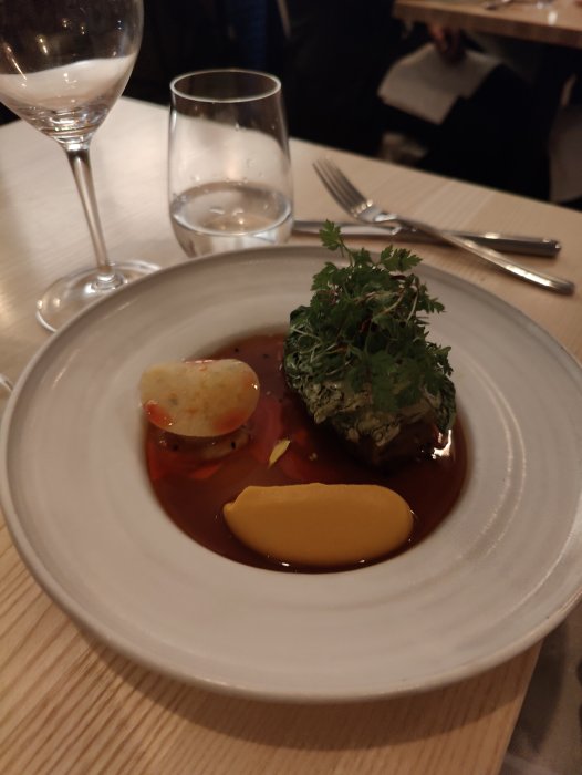 Elegant maträtt med gröna örter, beige sås, gastronomisk presentation, vin- och vattenglas, fint dukat bord.