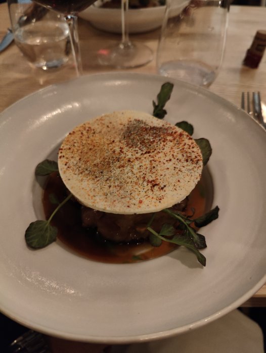 Gourmeträtt med rund kryddströdd skiva ovanpå, på en vit tallrik, omgiven av glas och bestick.