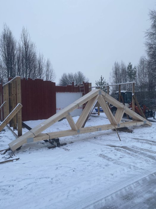 Byggplats med träkonstruktion i snö, oavslutat projekt, kallt landskap, grå himmel, ingen synlig aktivitet.