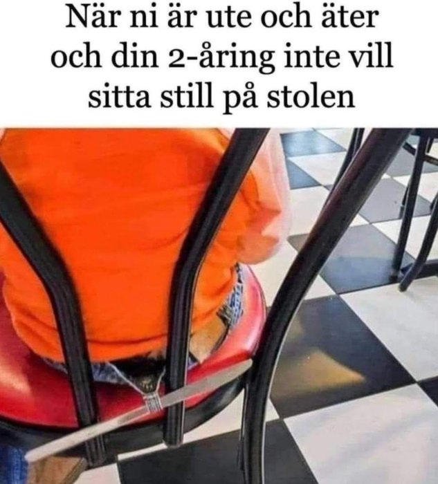 Barn klämt mellan stolsryggar, orange tröja, röd stol, svartvitt golv, humoristisk text, äta ute-tema.