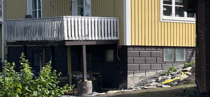 Hus med gul panel och balkong, stödben syns, byggnadsarbete nedanför, grönska i förgrunden.