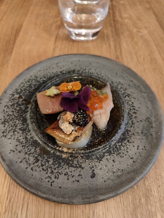 Delikat, konstnärlig sushirätt med blomma, fisk, kaviar och guldflarn på keramiktallrik, träbord i bakgrunden.