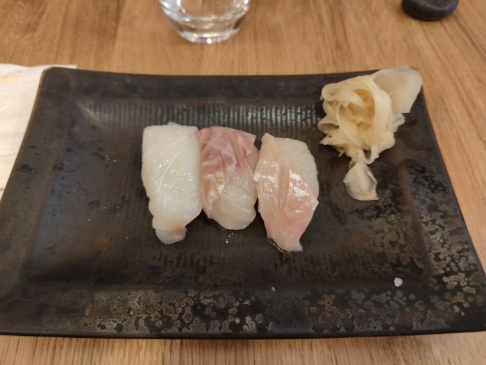 Tre bitar nigiri-sushi med vit fisk på en mörk keramiktallrik, garnerad med inlagd ingefära.