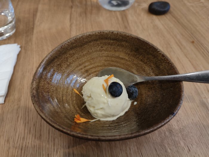 En skopa vaniljglass med blåbär och blomblad i keramikskål på träbord. Enkel, elegant dessertuppläggning.