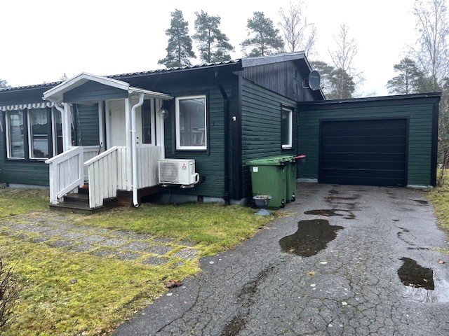 Enkelplanshus med veranda och garage, mulet väder, fuktig uppfart, grön soptunna, satellitantenn.