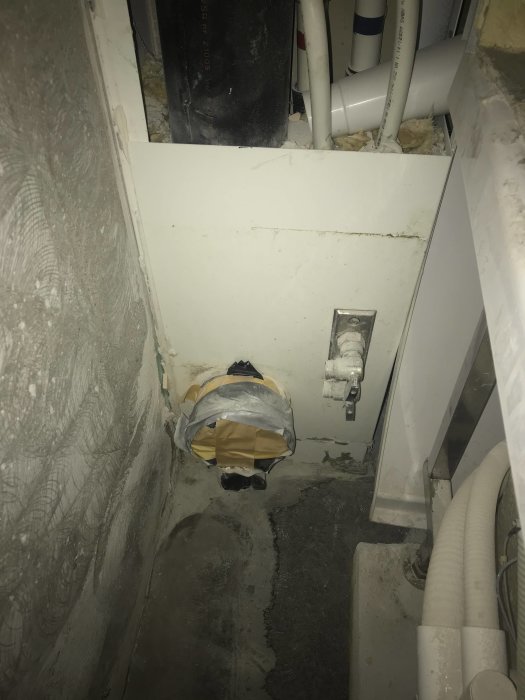 Gipsvägg med öppning, avskuren plastledning, betonggolv, rörinstallationer, slarvigt tejpade hörn, dolt utrymme.