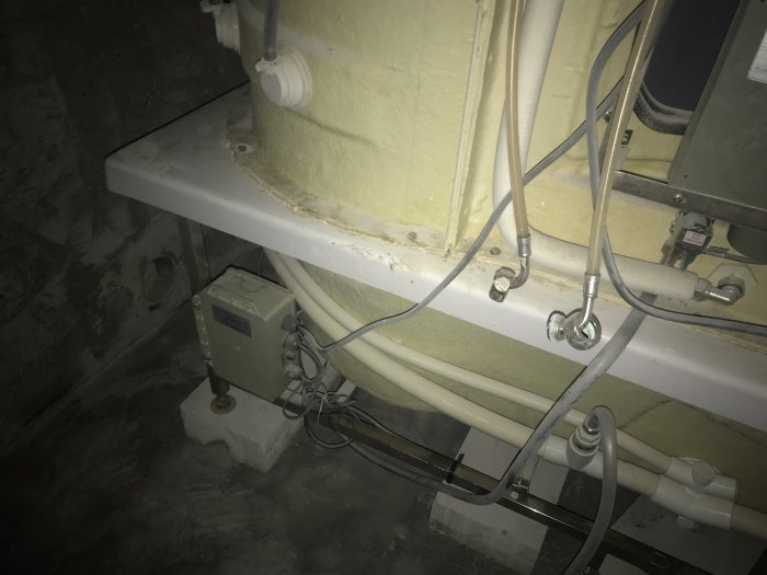 Industriell installation, kablar och rör, elektrisk utrustning, vit isolering, grå betonggolv, dålig belysning.