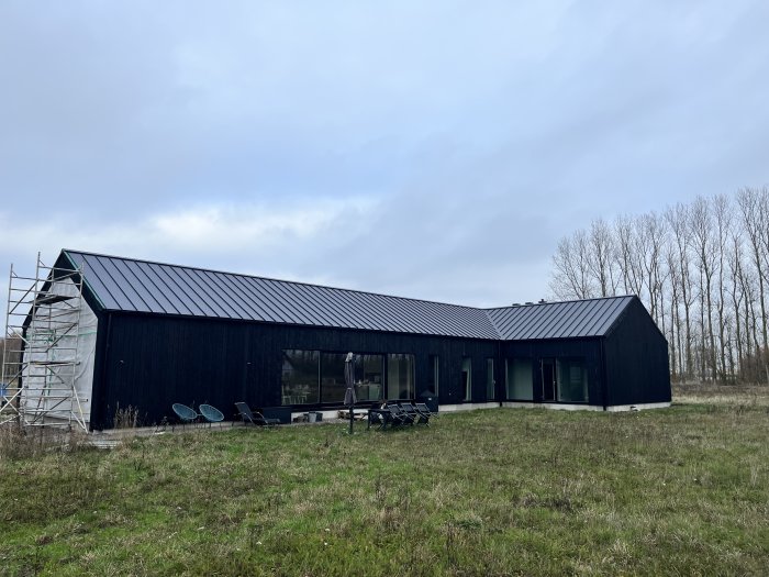 Svart hus med lutande tak, stora fönster, byggnadsställning, gräsmatta, träd i bakgrunden, molnig himmel.