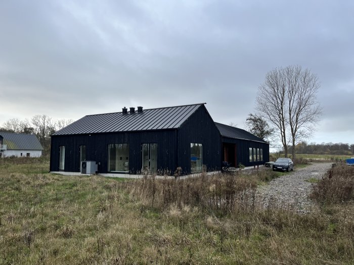 Modernt svart hus med stora fönster, grusväg och bil, omgivet av öppet landskap.