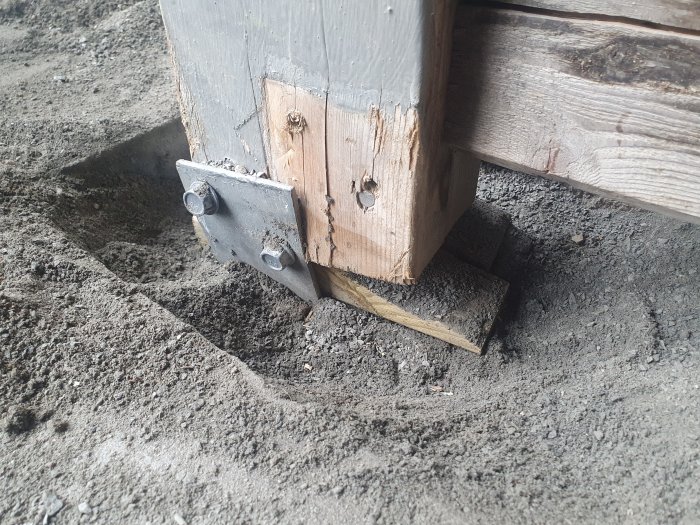 Träkonstruktion stabiliserad med metallbeslag och spik på grått, grovt underlag.