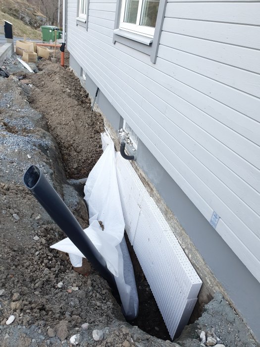 Grävd grund nära husvägg, dräneringsrör, isolermaterial, grus, byggarbetsplats, vit plast, utomhus.