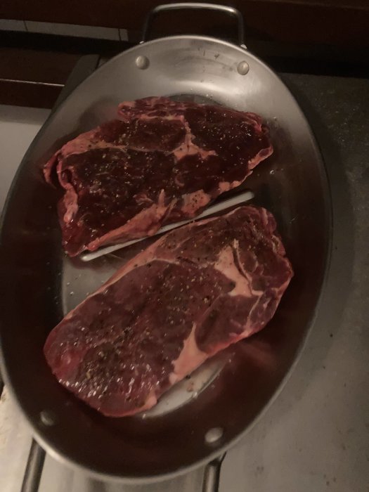 Två råa biffar kryddade på rostfritt stålplåt, redo för tillagning.
