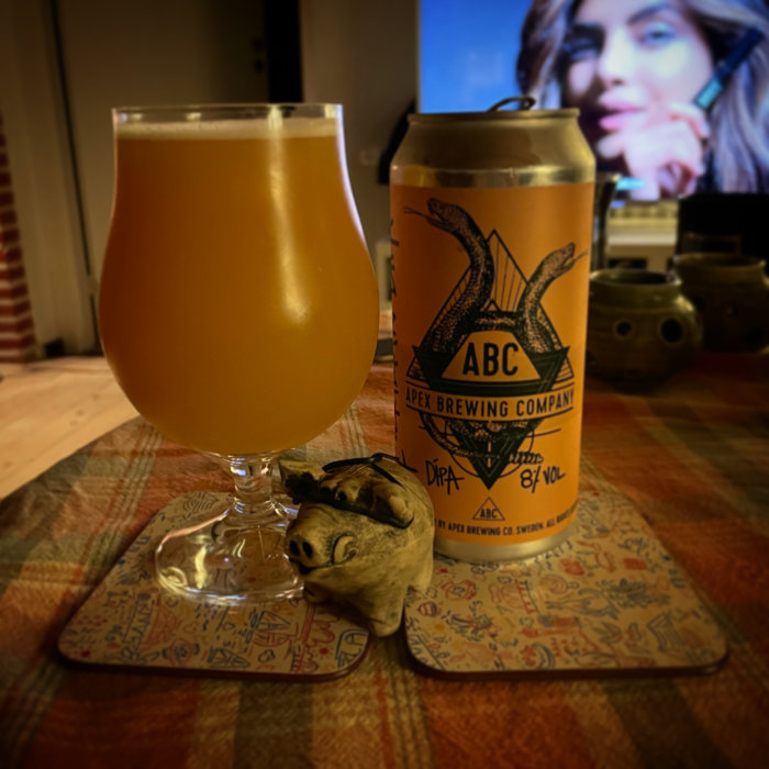 Ölglas med disig dryck, burk från Apex Brewing, underlägg, suddig bakgrund med person.