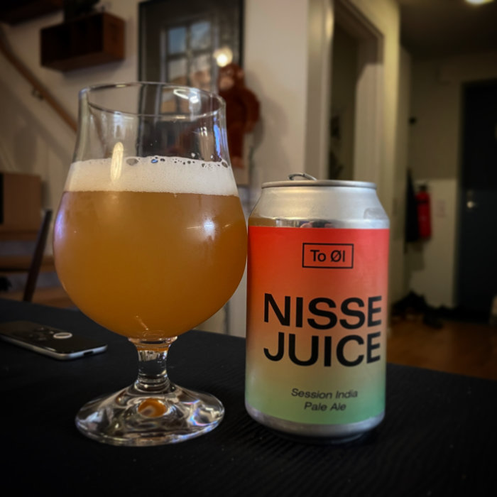 Ölglas med skum, burk märkt "NISSE JUICE Session India Pale Ale", inomhus, mysig atmosfär, suddig bakgrund.