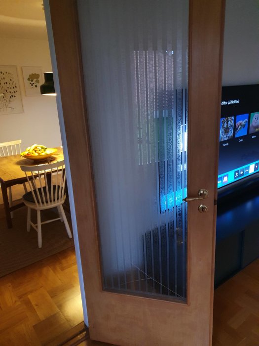 Halvöppen trädörr, frostat glas, inblick till vardagsrum, TV-skärm, Netflix-skärm, matbord, fruktskål, konst, hemmiljö.