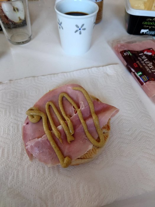 En smörgås med skinka och senap, kaffekopp och köttpaket på ett vitt pappersunderlägg.