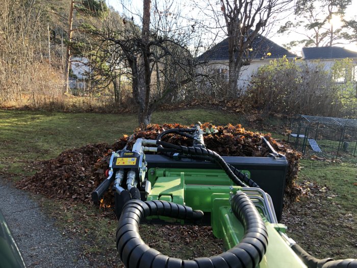 Trädgårdstraktor med lövuppsamlare fyllt med höstlöv framför tunt träd och hus.