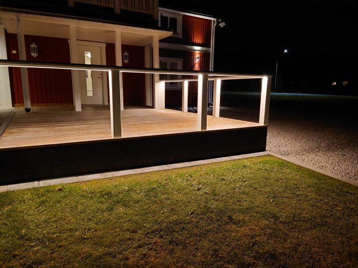 Veranda på kvällen, belysning, rött hus med vita knutar, gräs och gruset, inga människor syns.
