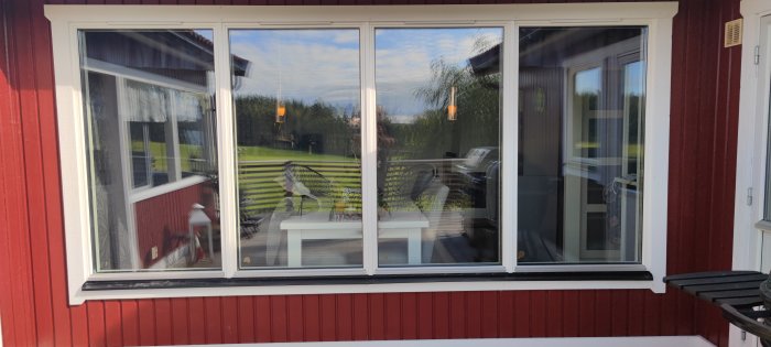 Stor fönster med spegelbild av trädgård, rött hus, uteplats och grill. Solljus och klar himmel.