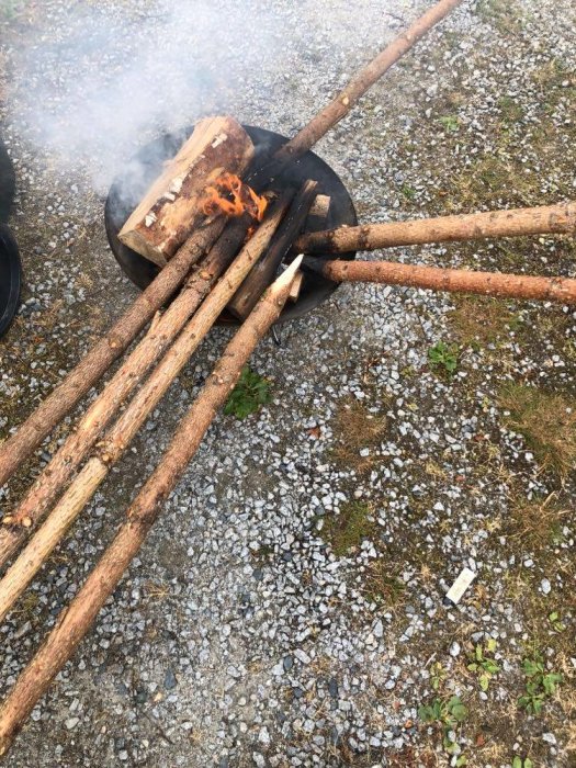 Eld i en utomhuspanna med rök, omgiven av långa pinnar, på grusig mark.