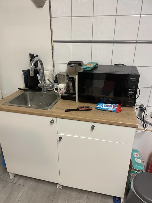 Litet köksutrymme med vask, mikrovågsugn, kaffebryggare, och personliga föremål. Vit kakelvägg och träbänk.