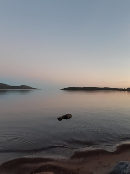 Skymning vid sjö, lugnt vatten, skog beklädda öar, flytande stock, rofylld himmel, sandstrand förgrund.