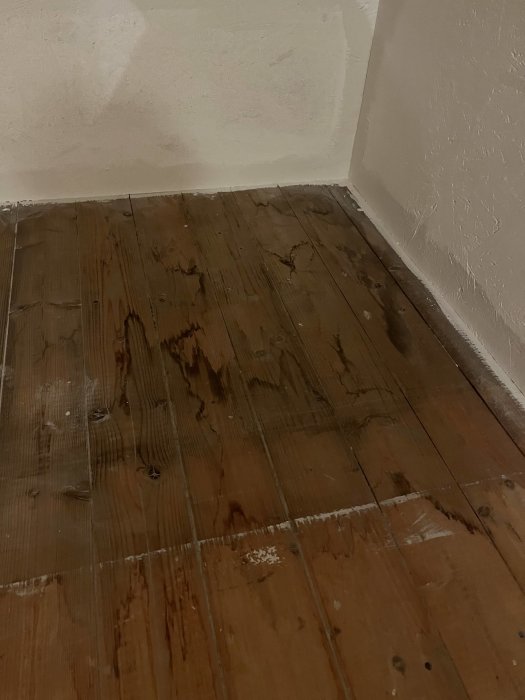Hörn av ett rum med mörkt trägolv och vitmålade väggar; fläckar och märken syns på golvytan.