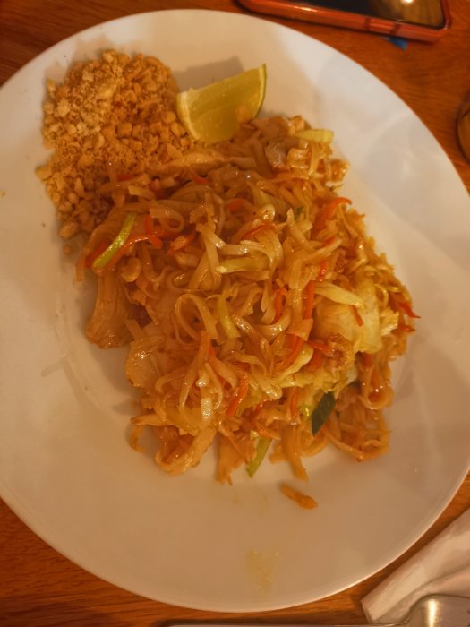 Pad thai med lime och jordnötter på en tallrik, på ett träbord, mobiltelefon i bakgrunden.
