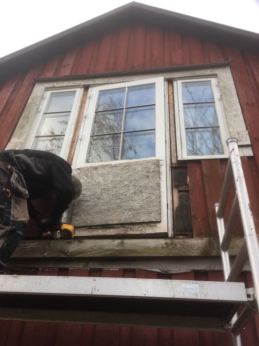 Person reparerar fönster på rödmålat hus, står på stegar, använder skruvdragare, molnig dag.