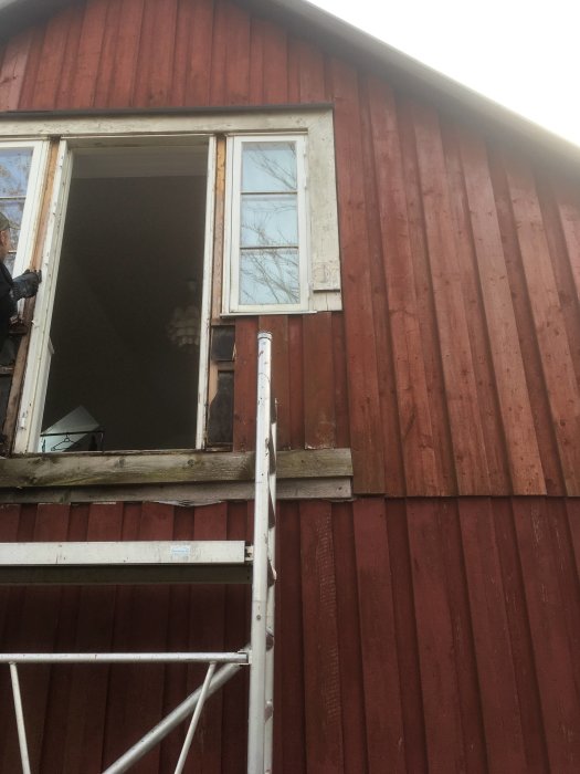 Rödfärgad trävägg, öppet fönster, stege, person i bakgrunden. Byggnadsrenovering eller fönsterbyte verkar pågå.