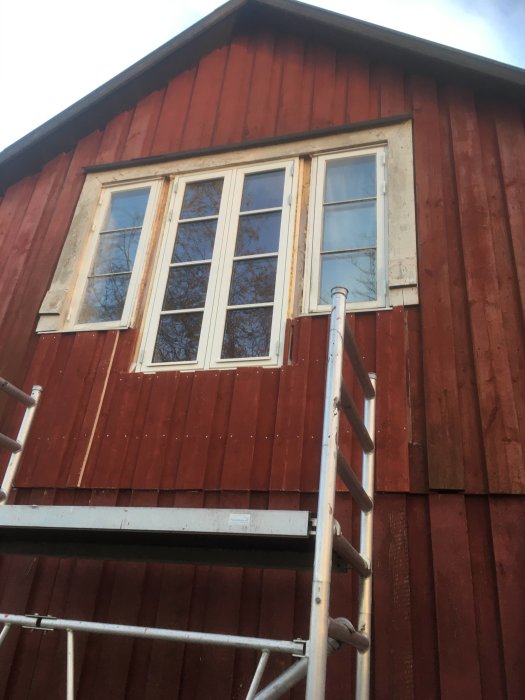 Röd stuga, två fönster, ställning, tegeltak i bakgrunden, dagtid, trolig renovering pågår.