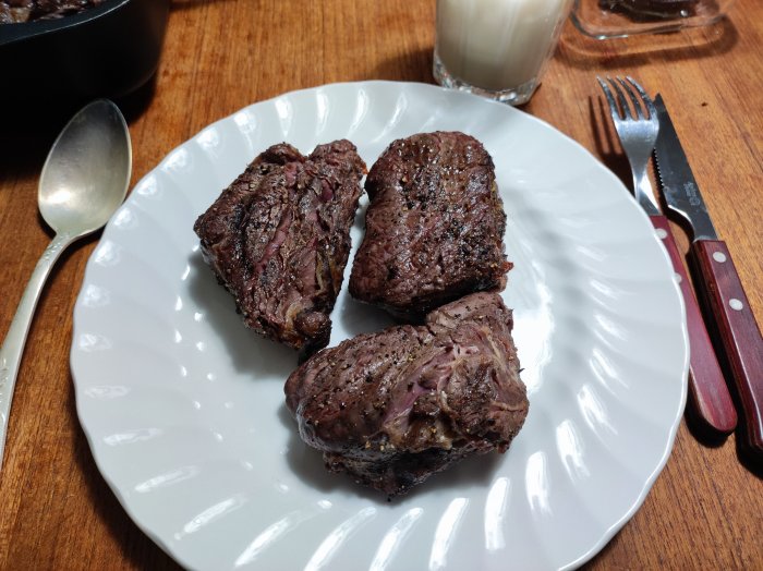 Tre grillade köttbitar på vit tallrik, träbordsbakgrund, bestick, sked och mjölkglas.