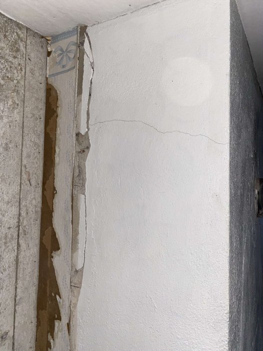 Skadad vägg med sprickor och avskalad gips eller färg, exponerat murverk och kakel. Behöver reparation eller renovering.